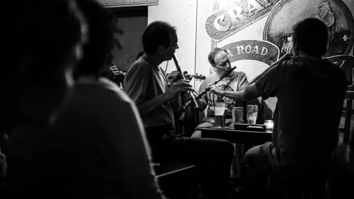 Black-and-white view of musicians seated around a pub table playing a whistle and a fiddle, with a large sign for a road on the wall behind them