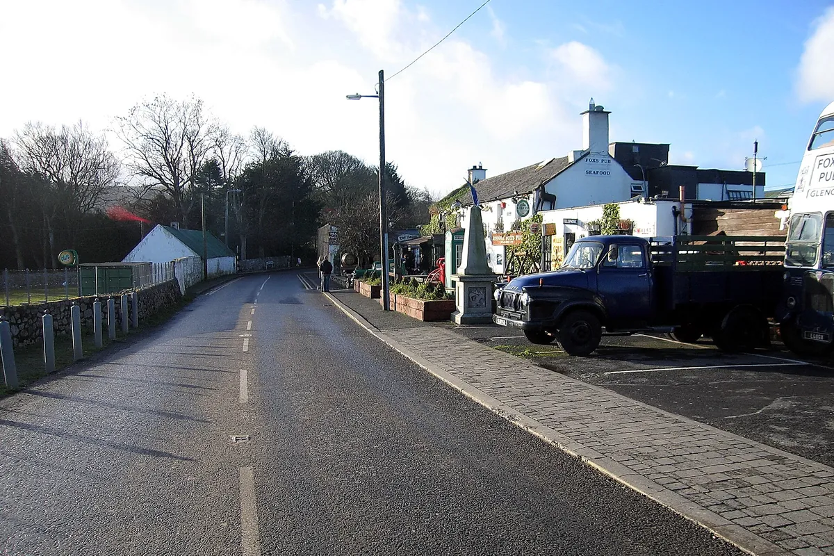 A quiet road through Glencullen village with a pavement on the right, bare trees beside it and the white buildings of Johnnie Fox's pub at the far end under a blue sky.