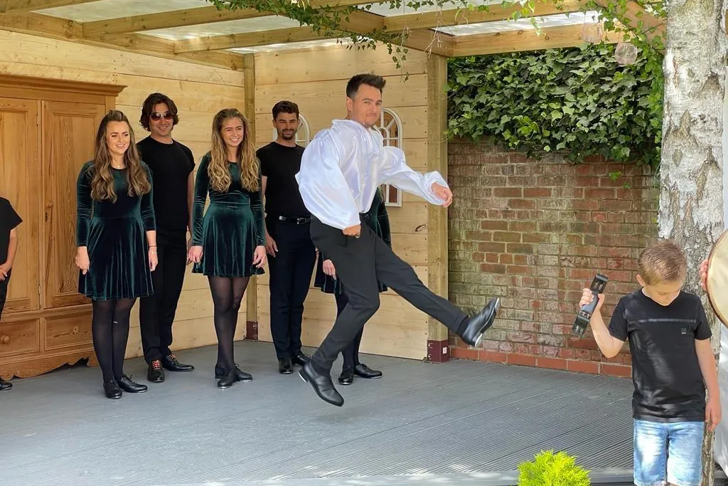 A male Irish dancer in a white shirt kicks one leg high on a grey wooden stage while a line of dancers in dark clothes stands behind him.