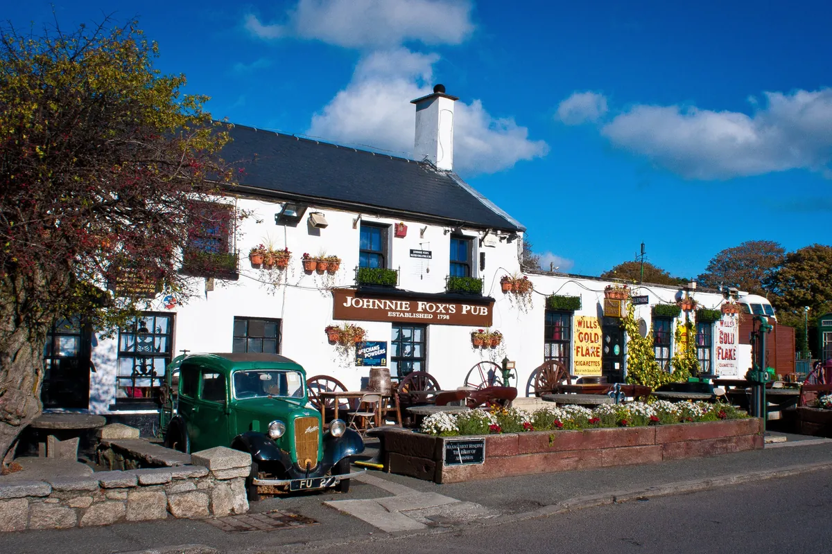 The white-painted front of Johnnie Fox's pub in Glencullen on a sunny day, with an old green car and flower beds outside and a dark slate roof above.