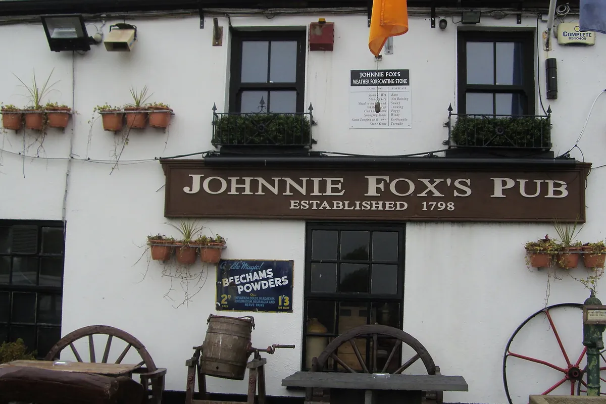 Whitewashed wall of a pub with a brown sign reading Johnnie Fox's Pub, Established 1798, flower pots on the wall and old cartwheels and a barrel outside