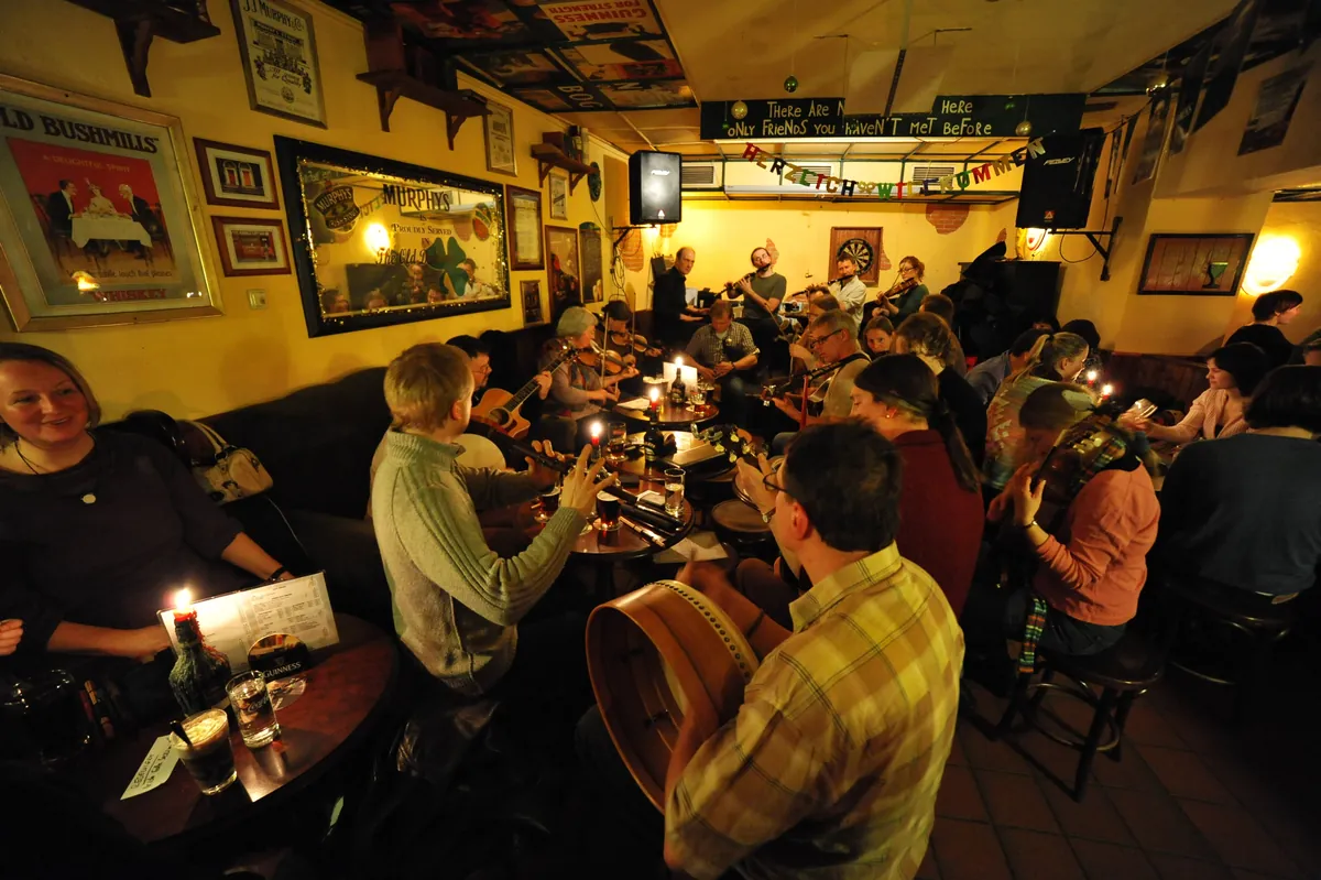 Musicians and guests crowd around candlelit tables at an Irish pub session, with a bodhrán drum in the foreground and old signs on the walls.